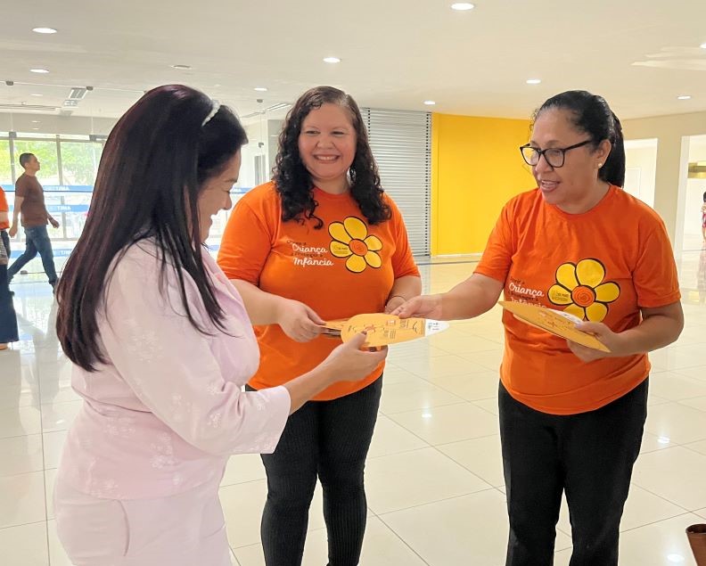 Duas servidoras do TJMA vestidas com camisas laranjas da campanha 18 de maio estão entregando panfletos para uma senhora no Fórum de São Luís.