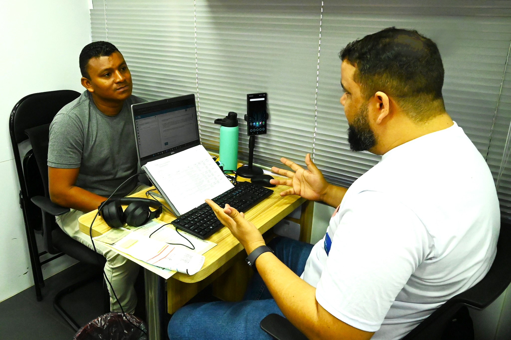 A imagem mostra duas pessoas sentadas em uma mesa em um ambiente de escritório. A pessoa à esquerda está de frente para um laptop e parece estar trabalhando, com papéis e um caderno sobre a mesa. A pessoa à direita está gesticulando com as mãos, possivelmente conversando ou explicando algo. Os rostos de ambas as pessoas estão ocultos para preservar a privacidade. Há itens pessoais, como garrafas de água, na mesa, e parece haver um quadro branco ou uma tela na parede atrás deles.