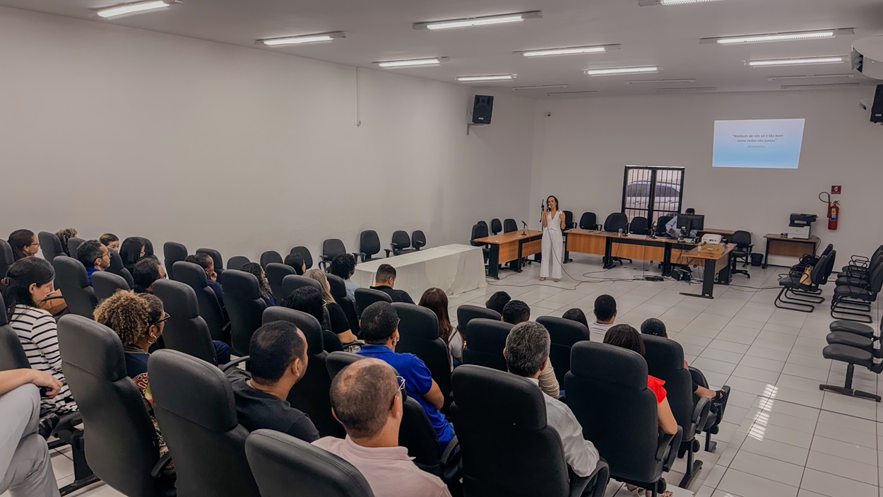 a imagem mostra um público de costas sentado em cadeiras pretas enquanto observam uma mulher vestida com roupas brancas falar ao telefone. ao fundo é possível observar cadeiras pretas e uma projeção de slides em parede branca