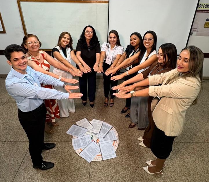 em ambiente interno, grupo de pessoas em pé dispostas em formato circular com os braços estendidos. no chao ao centro, uma série de objetos