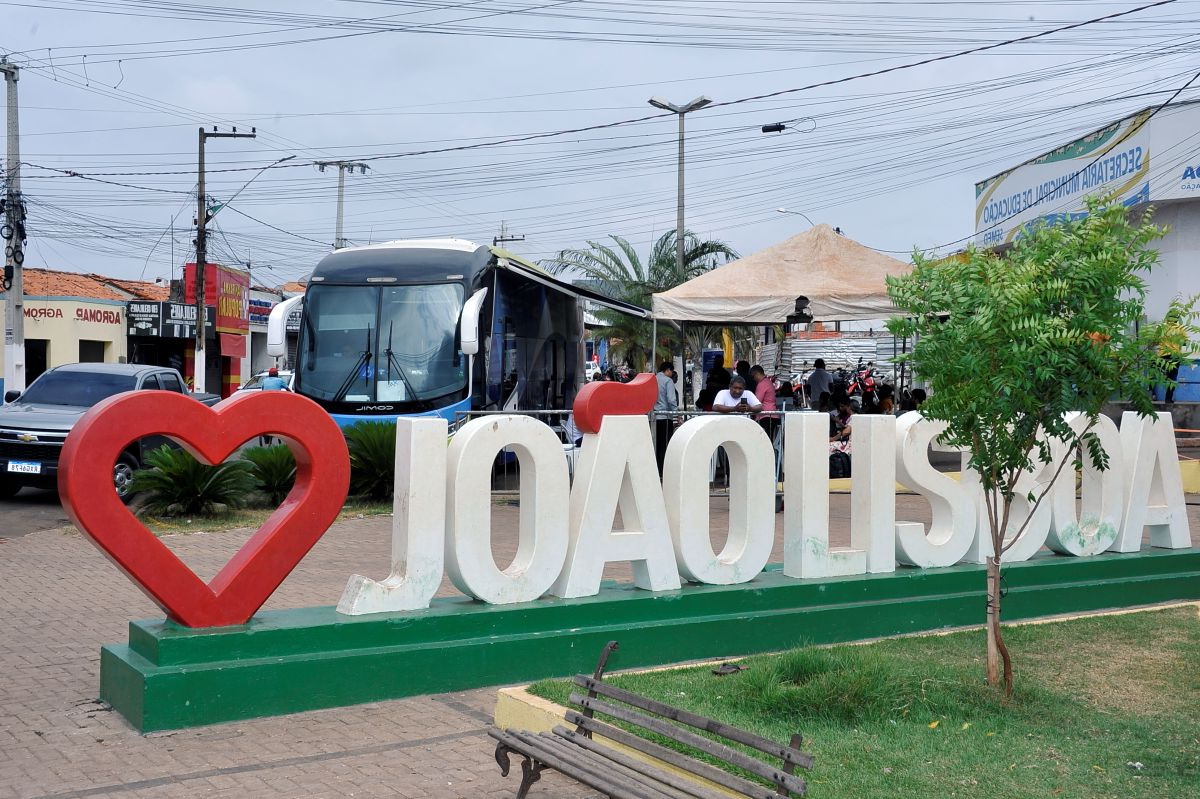 Foto horizontal. TJMA movimenta R$ 1,35 milhão em acordos na Região Tocantina. Essa imagem mostra a cidade de João Lisboa, no estado do Maranhão, Brasil. Em primeiro plano, há um letreiro grande e branco com as letras "JOÃO LISBOA" e, no lugar da última letra "A", um grande coração vermelho. Atrás do letreiro, é possível ver um banco de praça, uma pequena árvore e um espaço com barracas e pessoas. No lado direito, há um ônibus estacionado e prédios comerciais ao fundo.