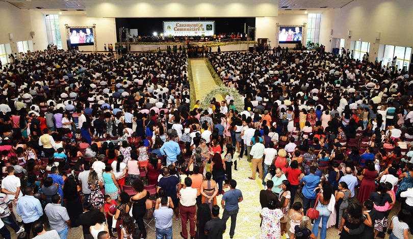 Imagem estatica de cima para baixo com centenas de pessoas em um espaço amplo, com banner ao fundo escrito casamento comunitário
