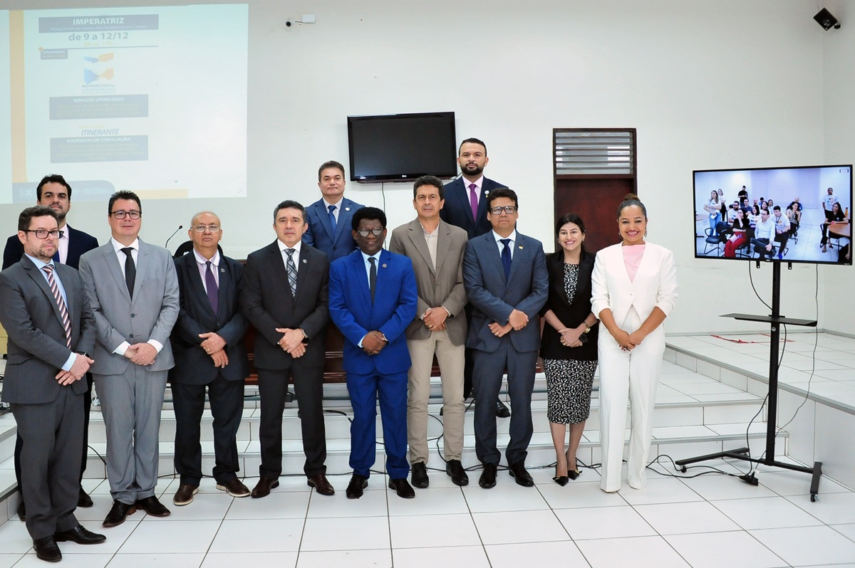 grupo de pessoas vestidas formalmente, em ambiente interno de auditório, em pé lado a lado. ao lado deles, uma televisão mostra aimagem de um outro grupo de pessoas em auditório, por videoconferência