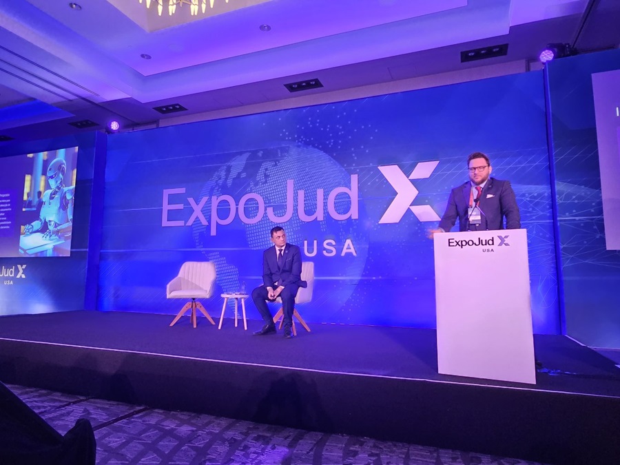 A imagem mostra um evento indoor com o juiz Felipe Damous como palestrante apresentando no palco, na frente de uma sala. A sala tem carpete azul e lustres modernos pendurados no teto. No palco, há uma grande tela exibindo um slide de uma apresentação com gráficos e texto relacionados ao ¿ExpoJud¿ e ¿Plataforma Digital do Judiciário.¿ O slide menciona ¿Interfaces: Sociedade de Automação.¿ Há banners em ambos os lados do palco com logotipos do ExpoJud USA.
