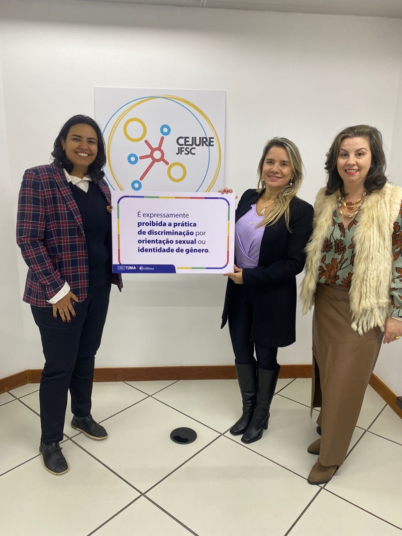 imagem mostra quatro pessoas em pé em frente a um banner com texto e gráficos. O banner apresenta um logotipo composto por círculos coloridos e linhas que se cruzam, lembrando um símbolo de átomo, com a sigla ¿CFJ/SC¿ abaixo. O texto no banner está em português e afirma que é expressamente proibida a discriminação por orientação sexual ou identidade de gênero.  Eles parecem estar posando para a fotografia, sugerindo que esta imagem pode estar relacionada a temas de inclusão, políticas anti-discriminação ou eventos que promovem esses valores dentro de uma organização ou instituição representada pela sigla no banner.