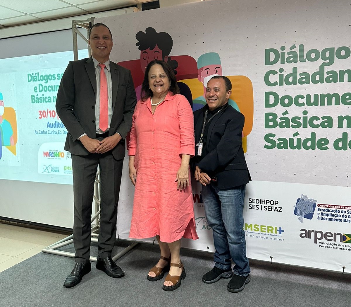 Dois homemns e uma mulher em pé, em frente a um banner do Encontro: Diálogos sobre Cidadania e Documentação Civil Básica na Rede de Saúde do Maranhão