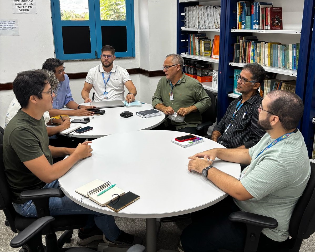 grupo de sete homens reunidos em torno de mesa em ambiente interno durante reuniao