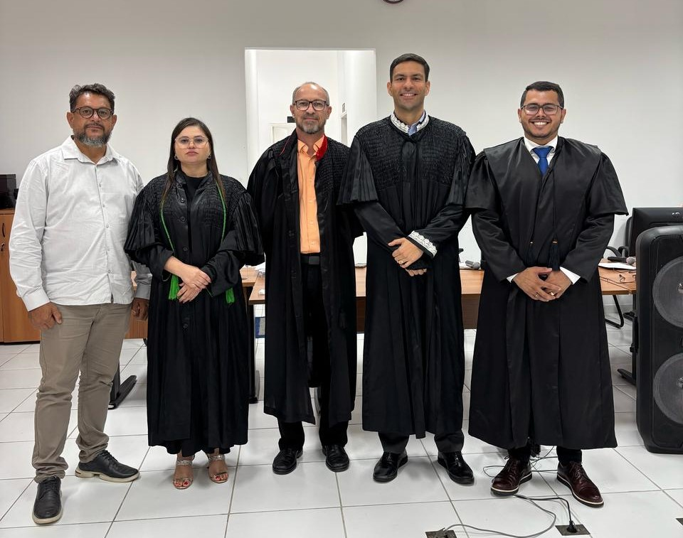 Foto horizontal, colorida, de três homens e uma mulher, vestidos de toga preta, de pé, em salão de teto e paredes brancas, posando pra foto.
