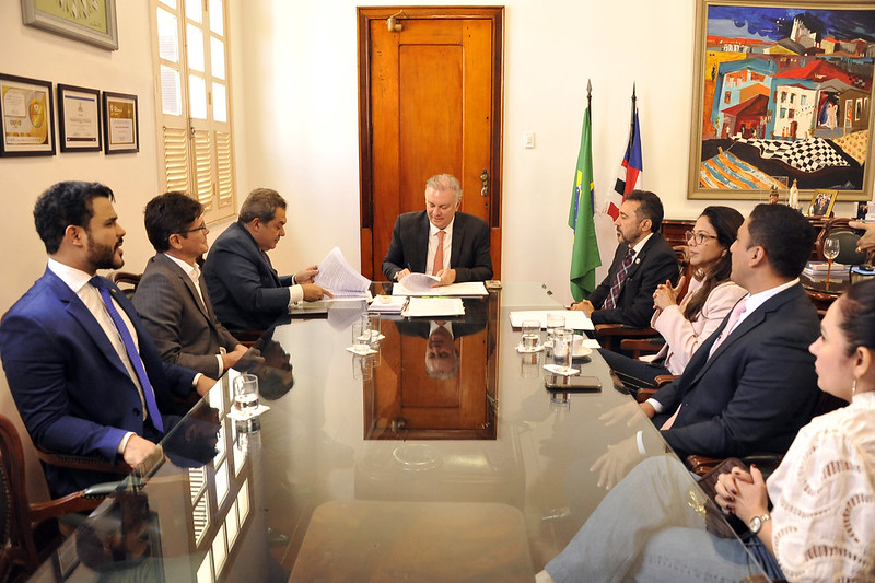 A imagem mostra um grupo de pessoas sentado em volta de uma grande mesa de conferência reflexiva em uma sala de reuniões formal. A pessoa na cabeceira da mesa está segurando um documento e parece estar liderando a reunião. Há vários documentos e copos de água sobre a mesa. A sala tem uma porta de madeira, certificados emoldurados na parede e uma pintura colorida. Duas bandeiras estão exibidas ao fundo, uma das quais é a bandeira do Brasil. O ambiente sugere uma reunião formal de negócios ou governamental.