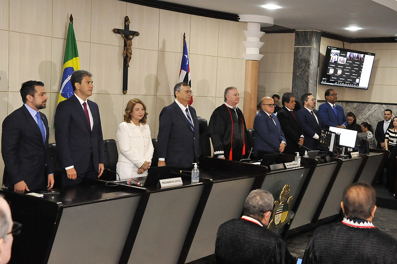 Foto horizontal da abertura do ano Judiciário 2025 do TJMA. A imagem mostra a cerimônia formal na sala das sessões plenárias do TJMA. Há várias pessoas em pé atrás de uma bancada, vestidas formalmente com ternos e togas. Na parede ao fundo, há uma cruz, a bandeira do Brasil e outra bandeira estadual. Há também uma placa na bancada com a inscrição "PROCURADOR DE JUSTIÇA". No canto superior direito, há uma tela exibindo uma videoconferência com várias janelas de participantes.