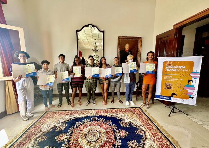 Foto horizontal. Entrega de novas certidões de nascimento à população trans, durante Mutirão Cidadania Transgênero. A imagem mostra um grupo de onze pessoas em pé, lado a lado, segurando certificados. Elas estão em um ambiente interno, com um espelho grande e um quadro na parede ao fundo. No chão, há um tapete grande e colorido. À direita da imagem, há um banner com o texto "CIDADANIA TRANSGÊNERO" e informações adicionais, incluindo um QR code. A imagem registra momento em que as pessoas estão recebendo certidões.