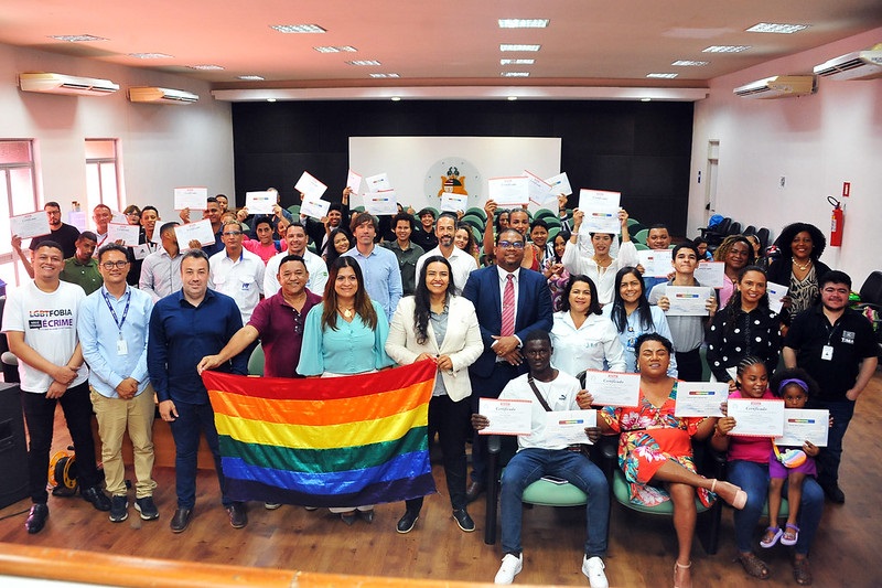 Foto horizontal. Homens e mulheres trans recebem certificação do curso de soldador - parceria com Montisol por Senai - mutirão trans. A imagem mostra um grupo grande de pessoas numa sala. As pessoas estão posando para uma foto em grupo, muitas delas segurando certificados. Na frente do grupo, há duas pessoas segurando uma bandeira do arco-íris, que é um símbolo do movimento LGBTQIA+. Algumas pessoas estão vestindo camisetas com mensagens, como uma que diz "LGBTFOBIA É CRIME". A sala tem um palco ao fundo com um pódio e um emblema na parede. A iluminação é fornecida por várias luzes de teto, e há aparelhos de ar-condicionado nas paredes laterais. A imagem é de um evento relacionado a direitos LGBTQIA+ e diversidade.