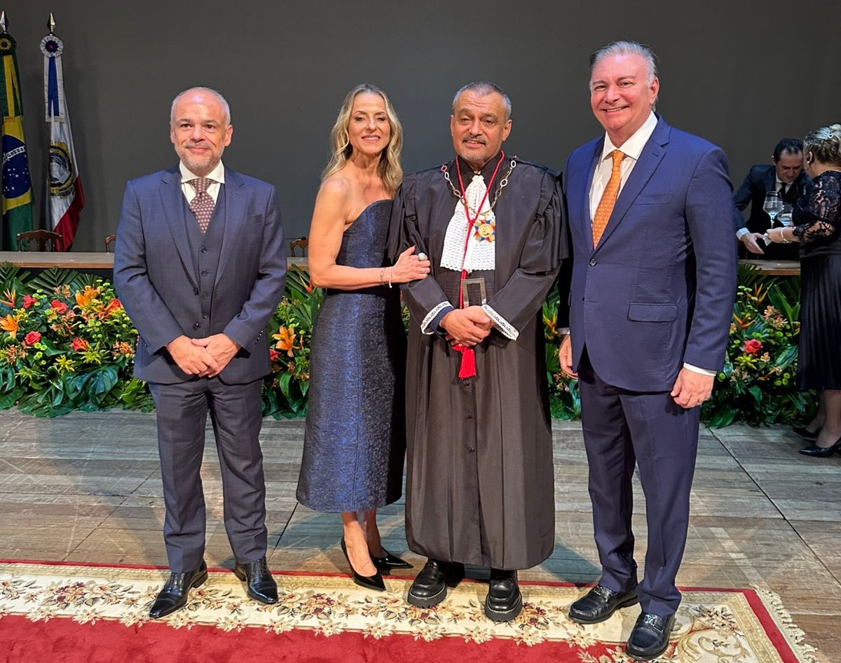 Foto horizontal da posse do presidente do TJAM. A imagem mostra quatro pessoas posando para uma foto, dentre elas o desembargador Froz Sobrinho, presidente do TJMA. Da esquerda para a direita, há um homem de terno cinza escuro, uma mulher de vestido azul escuro, o presidente do TJAM, desembargador Jomar Fernandes, usando uma toga preta e colar cerimonial, e o desembargador Froz Sobrinho, de terno azul escuro com gravata laranja. Ao fundo, há uma mesa com arranjos de flores coloridas e bandeiras, incluindo a bandeira do Brasil. O chão é coberto por um tapete com detalhes florais e borda vermelha.