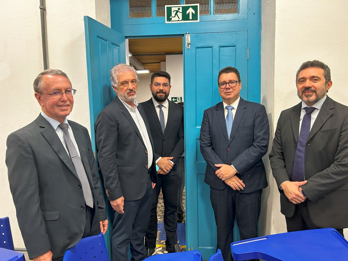 Foto horizontal - TJMA e UEMA definem local de Cejusc em Faculdade de Direito - Equipes Nupemec e UEMA. A imagem mostra cinco homens em pé, todos vestidos com ternos e gravatas, em frente a uma porta azul. A porta está aberta e há uma placa de saída de emergência verde com um pictograma de uma pessoa correndo acima da porta. O ambiente é um local interno, uma sala de aulas. Há cadeiras azuis visíveis na parte inferior da imagem.