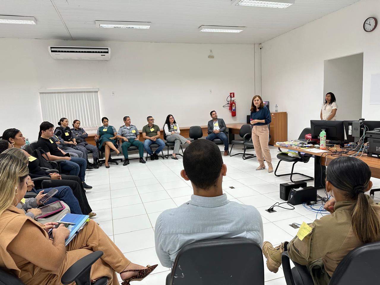 A imagem mostra um grupo de pessoas sentadas em um semi-círculo em uma sala, possivelmente participando de uma reunião ou workshop. A sala tem paredes brancas, piso de azulejos brancos e teto branco com lâmpadas fluorescentes. Há uma unidade de ar-condicionado montada na parede, um relógio e um extintor de incêndio. Alguns participantes estão usando uniformes, sugerindo que podem ser de uma organização ou profissão específica. Duas pessoas estão de pé na frente da sala, possivelmente conduzindo a sessão. Há mesas com computadores e vários itens, incluindo uma garrafa de água e alguns papéis. Os participantes parecem estar engajados, alguns tomando notas ou usando dispositivos eletrônicos.