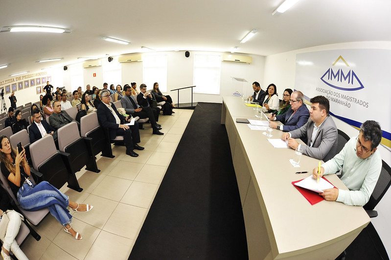 A imagem mostra uma reunião ou conferência formal acontecendo em uma sala bem iluminada. No lado direito da imagem, há um painel com seis indivíduos sentados atrás de uma mesa longa, cada um com documentos e materiais de escrita à sua frente. A parede atrás do painel exibe um logotipo com o texto "ASSOCIAÇÃO DOS MAGISTRADOS DO MARANHÃO." No lado esquerdo da imagem, há uma audiência sentada em filas de cadeiras, atenta ao painel. A audiência é composta por várias pessoas, algumas das quais estão fazendo anotações ou usando dispositivos eletrônicos. A sala tem uma atmosfera profissional, com foco na interação entre o painel e a audiência, indicando uma discussão ou apresentação formal.