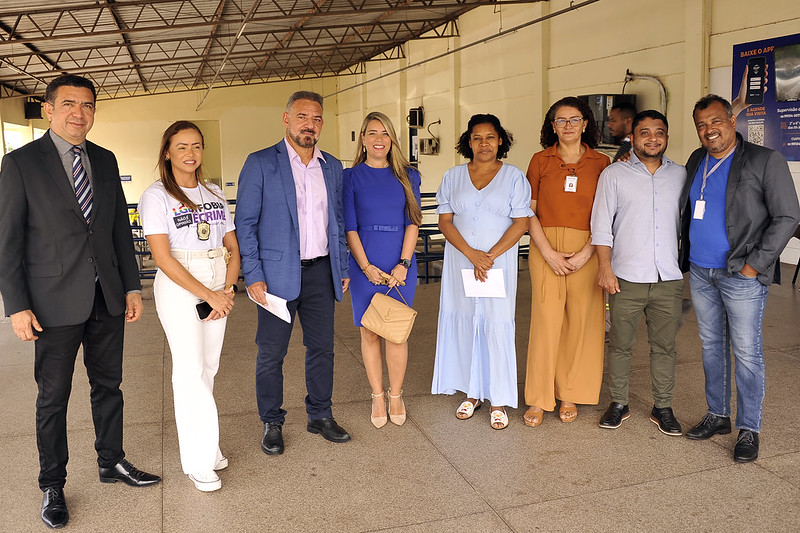 A imagem mostra um grupo de oito pessoas em pé, posando para uma foto. Os indivíduos estão vestidos com uma mistura de trajes formais e semi-formais, sugerindo um ambiente profissional ou oficial. O fundo inclui uma área coberta com um telhado de metal e alguns cartazes na parede. Um dos cartazes no lado direito da imagem tem um texto que diz "BAIXE O APP" e inclui um código QR, indicando uma promoção ou informação sobre o download de um aplicativo. As pessoas na imagem estão segurando papéis ou documentos, o que pode indicar que estão participando ou assistindo a um evento ou reunião.