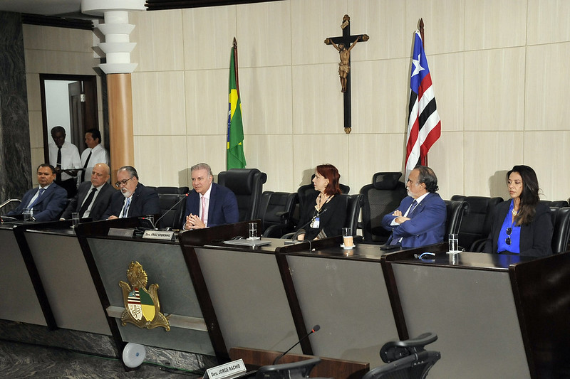 Imagem horizontal da Abertura da inspeção do CNJ no TJMA. A imagem mostra uma solenidade na sala das sessões plenárias do Tribunal. Há sete pessoas sentadas em uma mesa longa e curva, todas vestidas formalmente. Atrás da mesa, há duas bandeiras: a bandeira do Brasil, à esquerda, e a bandeira do Estado do Maranhão, à direita. No centro da parede, entre as bandeiras, há um crucifixo. Na frente da mesa, há um brasão de armas. As pessoas estão sentadas em cadeiras pretas e há microfones na mesa. Algumas pessoas estão olhando para a frente, enquanto outras estão conversando entre si. No fundo, à esquerda, há duas pessoas de pé perto de uma porta.