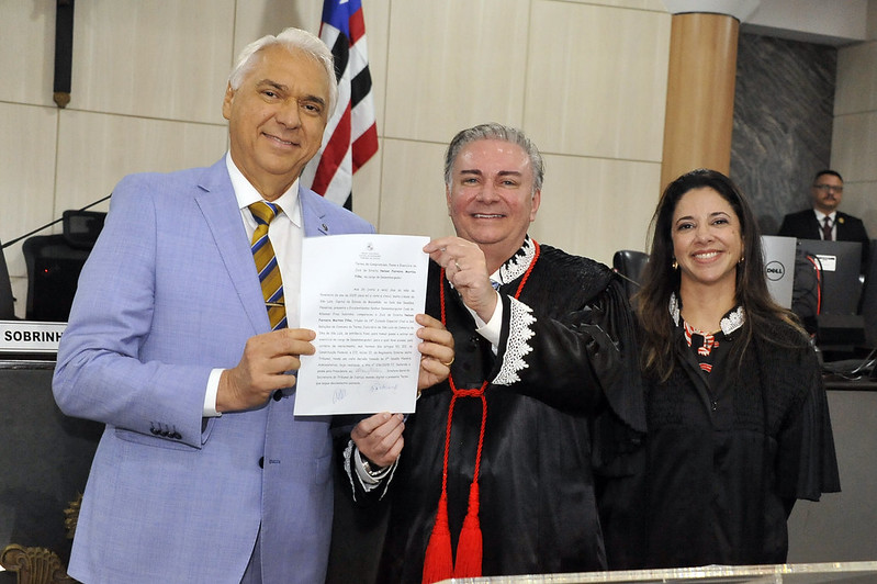 Foto horizontal de evento de eleição e posse do desembargador Nélson Ferreira. A imagem mostra três pessoas em pé. Duas delas, o empossado e o presidente do TJMA, desembargador Froz Sobrinho, estão segurando o termo de posse. Nélson, à esquerda, está vestido com um terno azul claro e uma gravata listrada. Froz Sobrinho, ao centro, veste uma toga preta com detalhes vermelhos e brancos, e a pessoa à direita, a diretora do TJMA, juíza Ticiany Palácio, também está vestida com uma toga preta. O documento que está sendo segurado contém texto, mas não é possível ler todo o conteúdo devido à resolução da imagem. Ao fundo, há uma bandeira com listras verticais nas cores preta, branca e vermelha, do Maranhão.