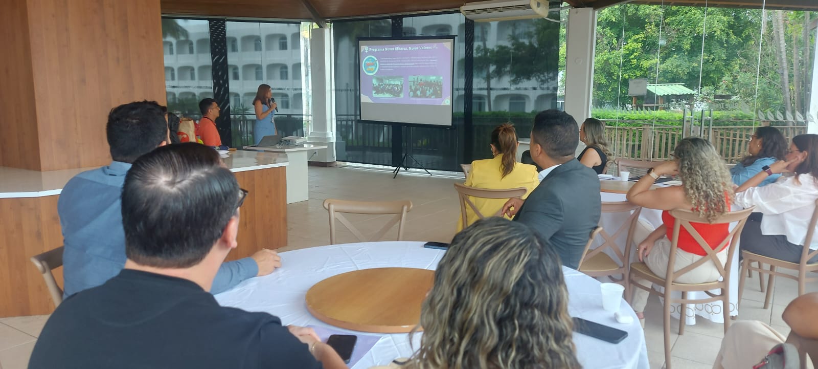 A imagem mostra um grupo de pessoas sentadas ao redor de uma mesa em um ambiente semiaberto, participando de uma apresentação. O apresentador está em pé perto de uma tela exibindo um slide que inclui texto e imagens, mas os detalhes não estão totalmente legíveis. O ambiente parece ser profissional ou educacional, com os participantes prestando atenção à apresentação. A sala tem grandes janelas com vista para uma área verde do lado de fora. A imagem captura um momento de aprendizado ou compartilhamento de informações em um ambiente formal.