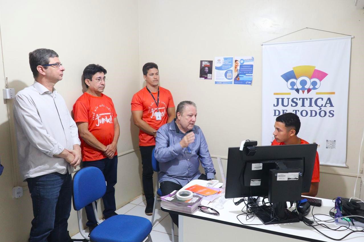 A imagem mostra cinco pessoas em um ambiente de trabalho ou atendimento. Três delas estão em pé, vestindo camisetas vermelhas com a palavra "Juntos!" escrita na frente. Uma pessoa está sentada em uma cadeira azul, usando uma camisa azul e gesticulando com a mão direita. Outra pessoa está sentada em frente a um computador, também vestindo uma camiseta vermelha. Ao fundo, há um banner com o texto "JUSTIÇA DE TODOS" e alguns cartazes menores na parede. A sala é equipada com cadeiras azuis e uma mesa branca com alguns papéis e objetos sobre ela. Parece ser um espaço dedicado à prestação de serviços ou suporte.