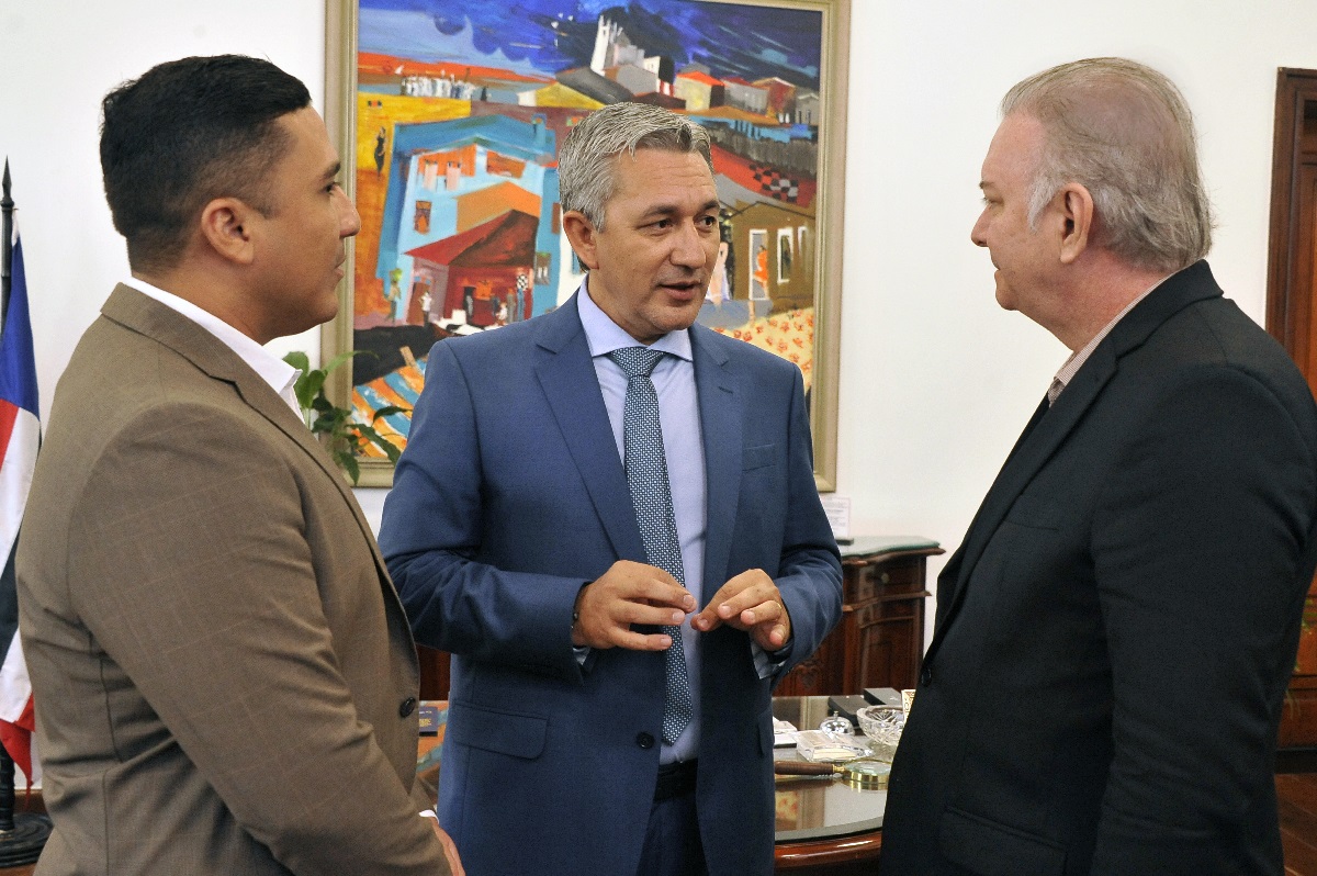 Foto horizontal da visita do novo presidente da Arpen-Brasil visita TJMA. A imagem mostra três homens em pé, conversando em um ambiente interno. O homem à esquerda está vestindo um terno marrom claro com uma camisa branca. O homem no centro está vestindo um terno azul com uma camisa azul clara e uma gravata azul com padrões. O homem à direita está vestindo um terno preto com uma camisa clara. Ao fundo, há uma pintura colorida com várias casas e uma paisagem urbana, além de uma bandeira parcialmente visível à esquerda.
