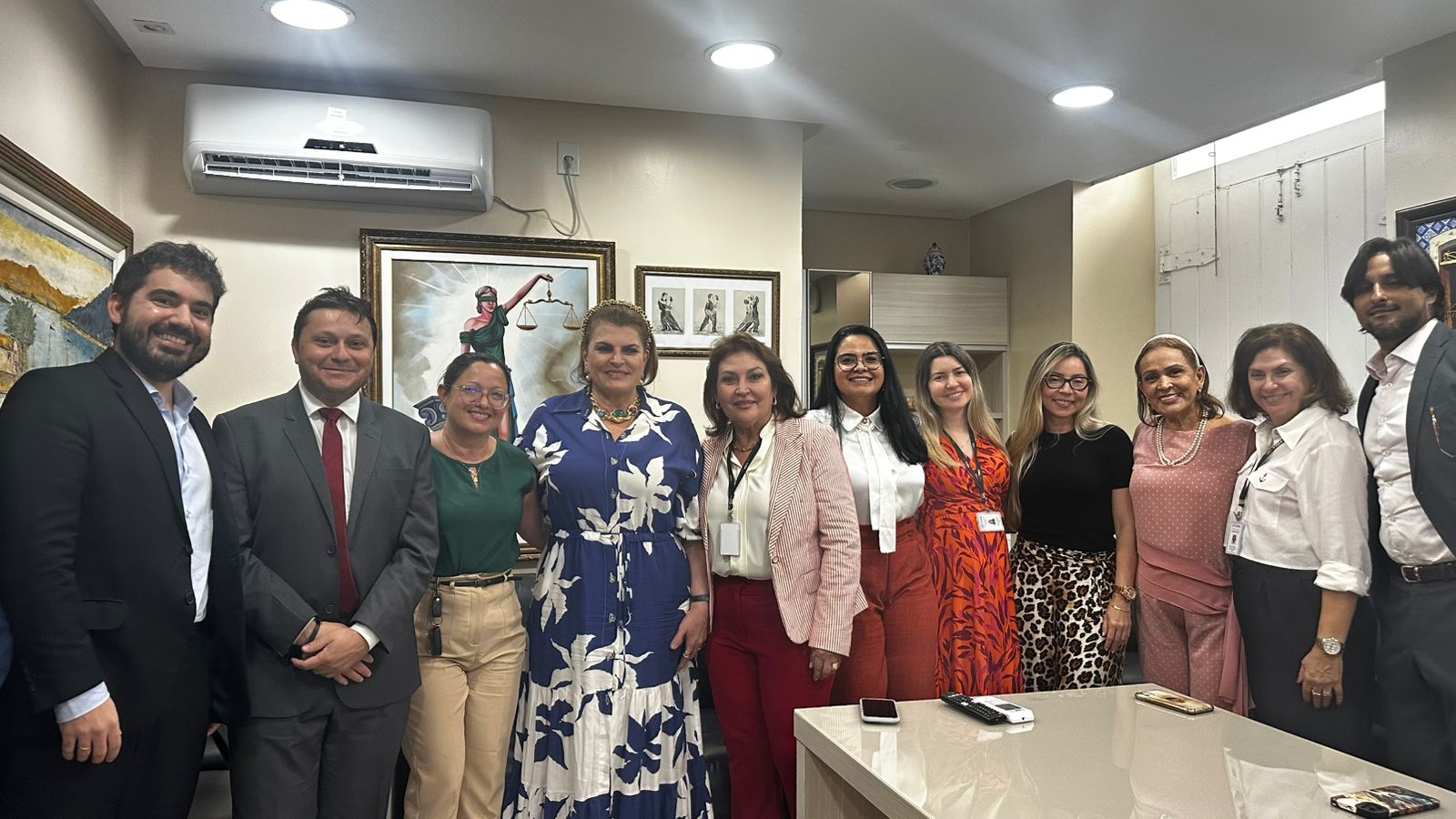 A imagem mostra um grupo de pessoas reunidas em um ambiente de escritório. As pessoas estão vestidas com roupas formais e semi-formais, sugerindo um cenário profissional. No fundo, há várias obras de arte, incluindo uma pintura proeminente da Dama da Justiça segurando uma balança, frequentemente associada a ambientes jurídicos.Há também uma unidade de ar-condicionado montada na parede acima do grupo. A sala é bem iluminada com luzes no teto e há uma mesa em primeiro plano com um controle remoto e um smartphone sobre ela.