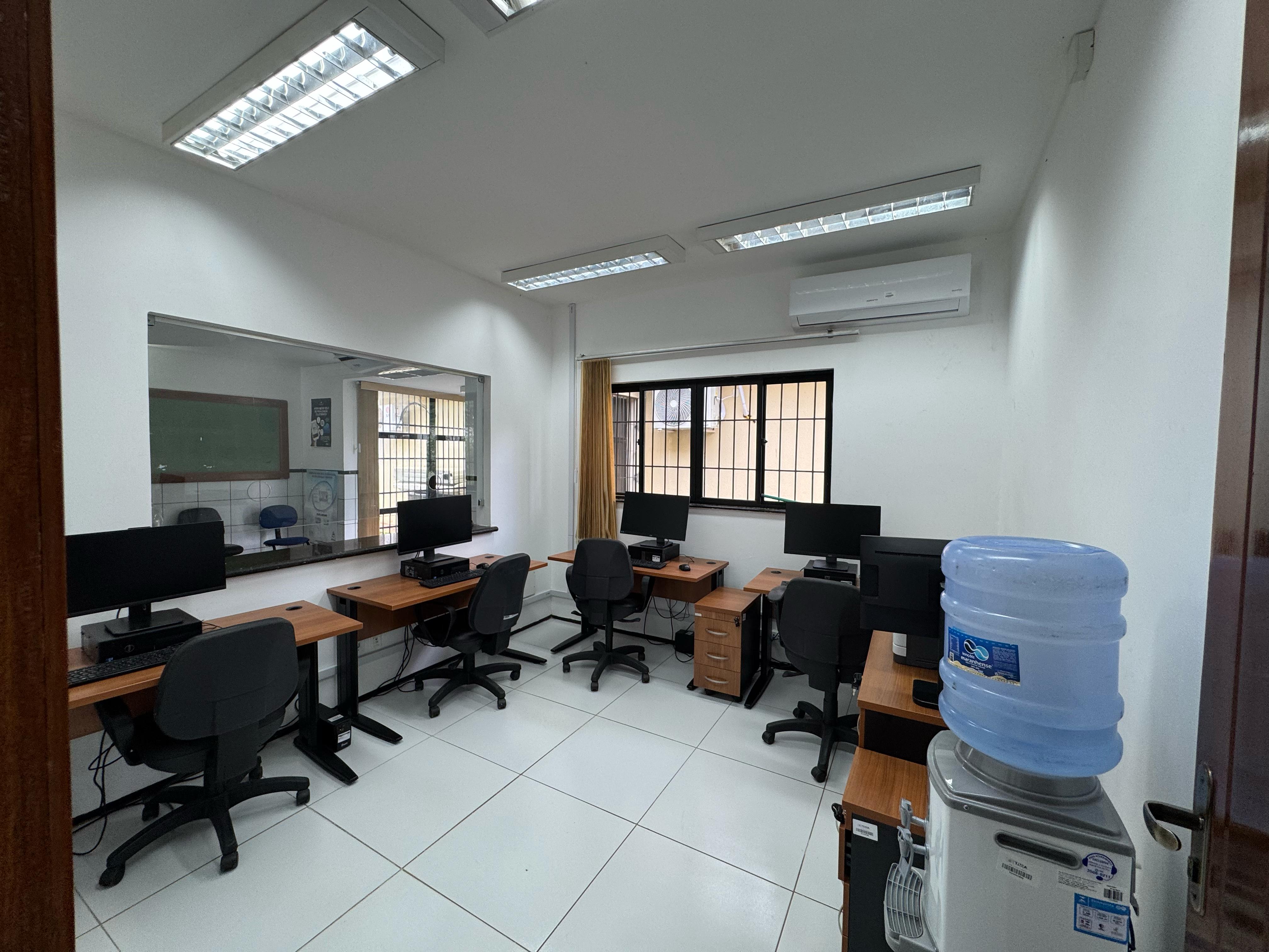 A imagem mostra uma sala de informática com várias estações de trabalho. Há cinco mesas de madeira, cada uma equipada com um computador de mesa e uma cadeira de escritório preta. As mesas estão dispostas ao longo das paredes da sala. No canto direito da imagem, há um bebedouro com um garrafão de água azul. A sala é iluminada por luzes fluorescentes no teto e possui um aparelho de ar-condicionado instalado na parede.
