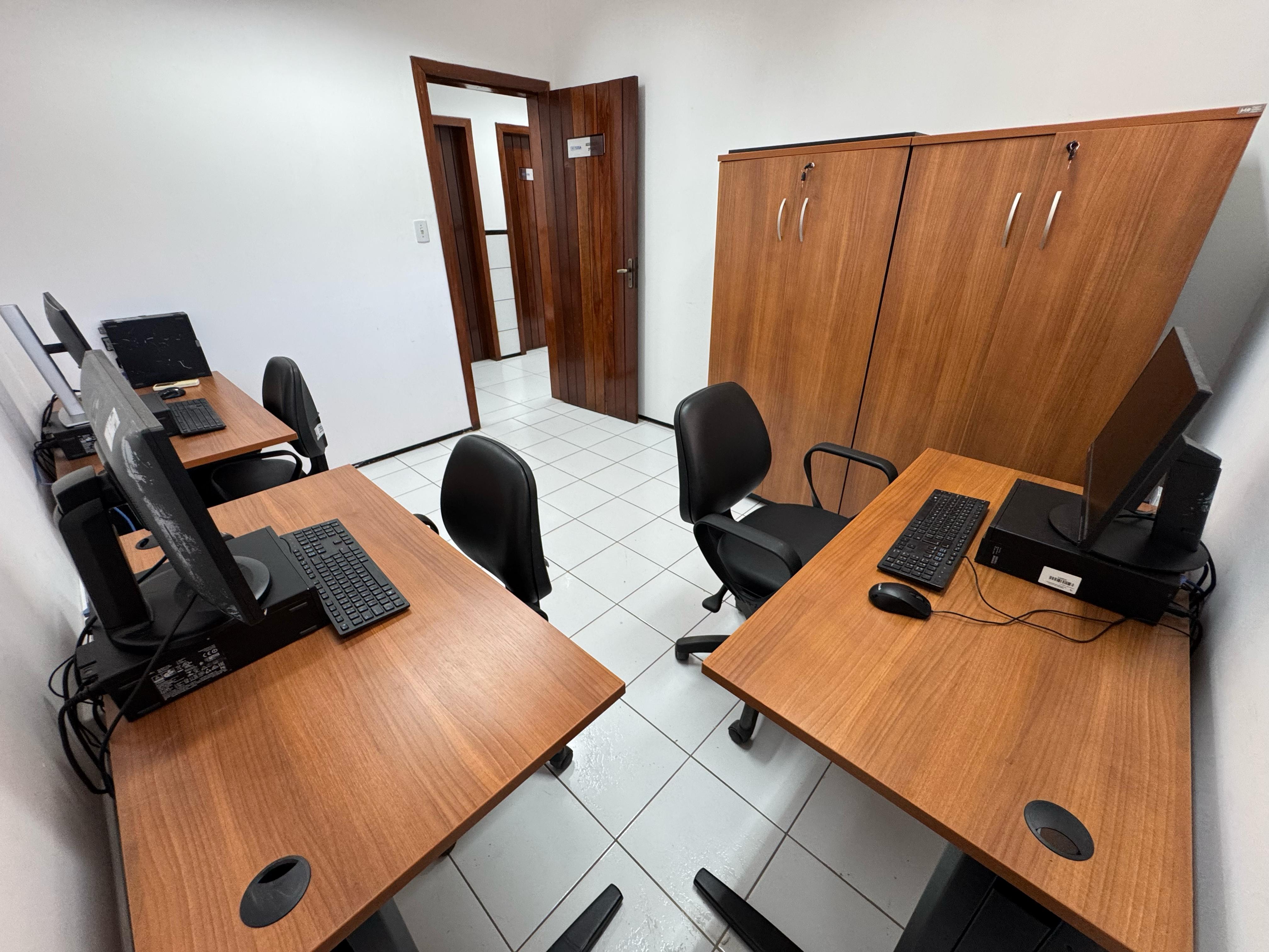 A imagem mostra uma sala de informática com quatro estações de trabalho. Cada estação possui uma mesa de madeira com um computador de mesa, teclado e mouse. As cadeiras são pretas e giratórias. No fundo da sala, há dois armários de madeira com portas fechadas. A sala tem piso de cerâmica branca e paredes brancas. Há uma porta de madeira aberta que leva a um corredor.