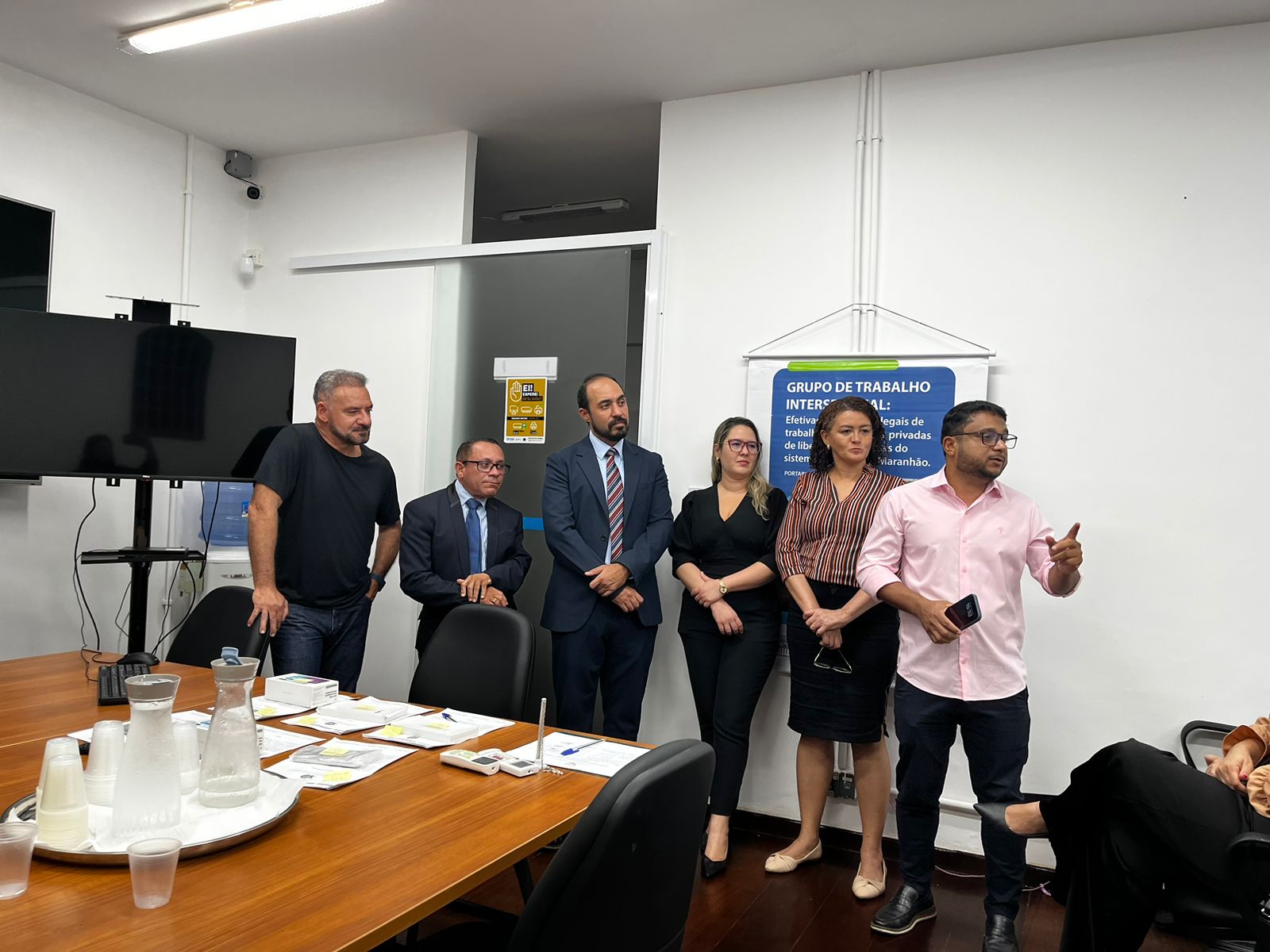 A imagem mostra um grupo de seis pessoas em pé em uma sala de reunião. Há uma mesa de madeira com vários documentos, garrafas de água e copos descartáveis sobre ela. Na parede ao fundo, há um cartaz com o título "GRUPO DE TRABALHO INTERSETORIAL" e um texto explicativo abaixo. Ao lado do cartaz, há uma porta com um aviso amarelo. À esquerda, há uma televisão de tela plana montada na parede. A sala é iluminada por luzes fluorescentes no teto.