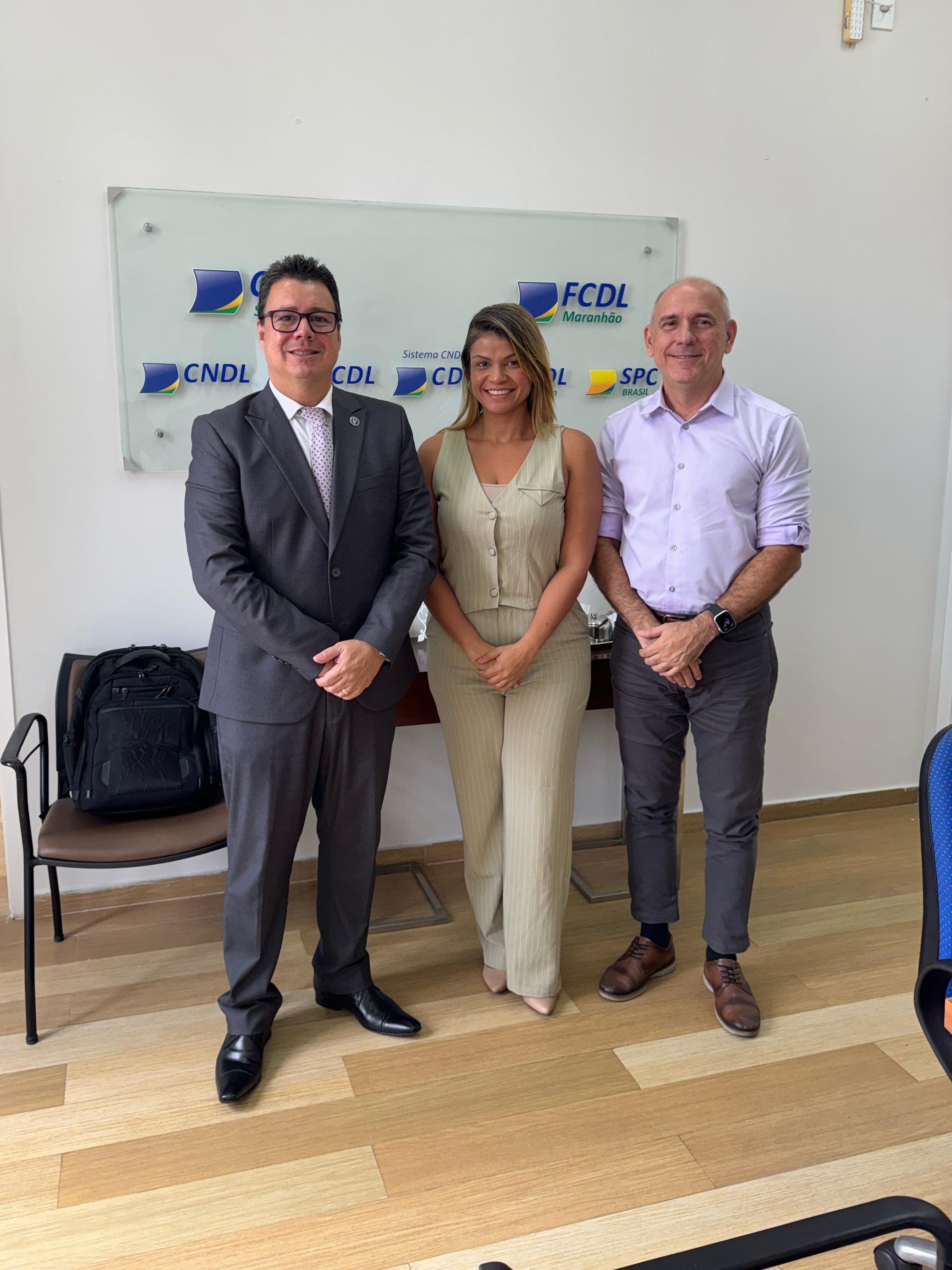 A imagem mostra uma reunião de negócios em uma sala de conferências. Quatro pessoas estão sentadas ao redor de uma mesa de madeira, discutindo ou trabalhando em algo. Na mesa, há vários itens, incluindo laptops, um calendário de mesa com o ano 2025, uma garrafa de água, um telefone de conferência e outros materiais de escritório.

Na parede ao fundo, há uma placa com os logotipos CDL São Luís, FCDL Maranhão, CNDL, FCDL, CDL e SPC. A sala é bem iluminada, com uma porta aberta à esquerda e uma janela com persianas à direita.