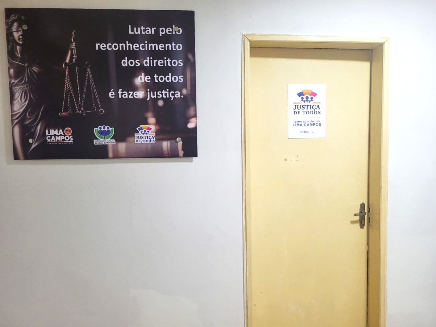 A imagem mostra duas pessoas sentadas em uma mesa, vestindo trajes formais com detalhes em renda branca e cordões vermelhos ao redor do pescoço. Ambas as pessoas estão segurando papéis, e uma delas está falando em um microfone. Há um laptop e mais papéis sobre a mesa. Ao fundo, outras pessoas também estão presentes, sugerindo um ambiente formal, possivelmente uma reunião ou sessão de tribunal.