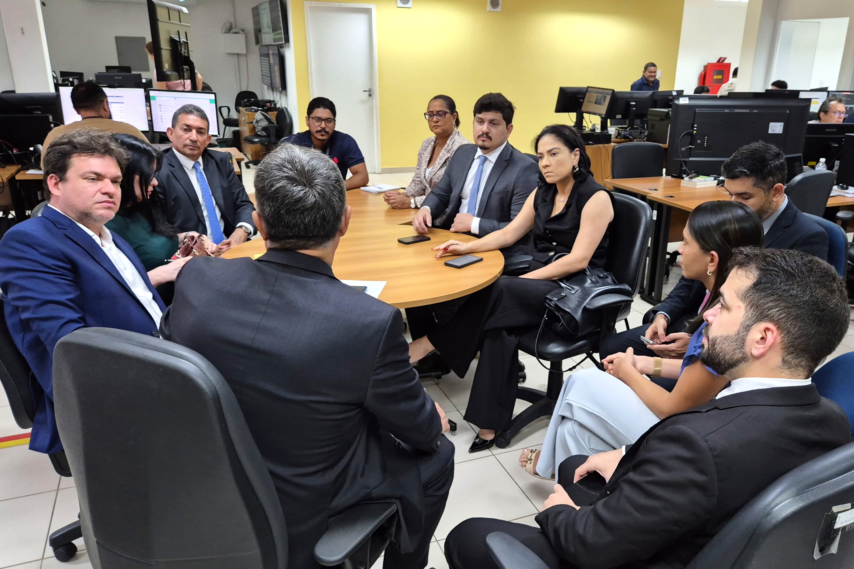 Claro! A imagem mostra um grupo de pessoas em uma reunião em um escritório. As pessoas estão sentadas em cadeiras ao redor de uma mesa, algumas delas usando ternos e outras roupas formais. Ao fundo, há várias mesas de trabalho com computadores e monitores, onde outras pessoas estão trabalhando. A sala é bem iluminada, com luzes fluorescentes no teto e grandes janelas que permitem a entrada de luz natural. A imagem é interessante porque mostra um ambiente de trabalho colaborativo, onde várias pessoas estão envolvidas em uma discussão ou reunião, enquanto outras continuam suas atividades em suas estações de trabalho
