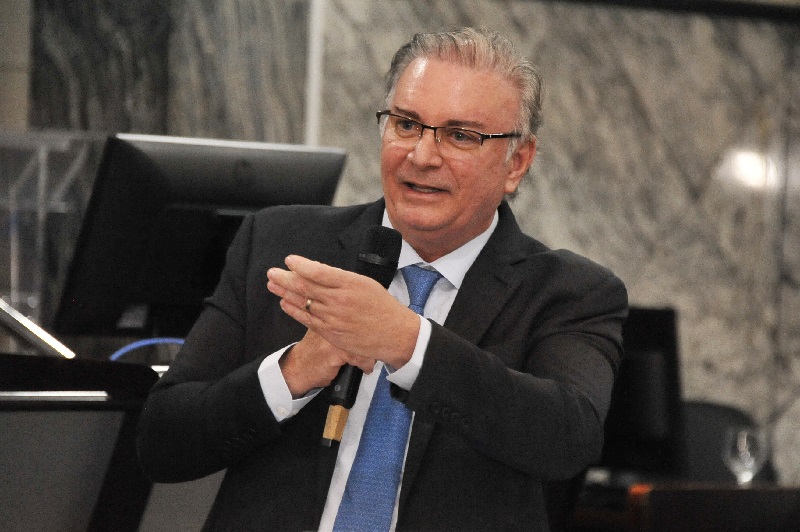 Foto horizontal do desembargador Froz Sobrinho, presidente do Tribunal de Justiça do Maranhão. A imagem mostra o desembargador, homem branco, meia idade, cabelos grisalhos, vestindo terno escuro, camisa branca e gravata azul. Ele segura um microfone com uma das mãos enquanto gesticula com a outra. Ao fundo, há um monitor de computador e uma parede com um padrão de mármore, na Sala das Sessões Plenárias do Tribunal.