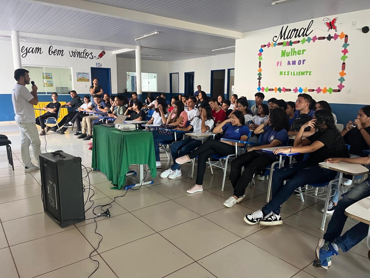 Na terceira imagem, o palestrante aparece em pé, segurando um microfone e falando para os alunos. Ele veste roupas claras e está ao lado de uma mesa coberta com toalha verde, sobre a qual há um projetor. Em frente a ele, uma caixa de som grande está posicionada no chão. Os estudantes, sentados, escutam com atenção. Ao fundo, há mais murais nas paredes com frases como ¿Sejam bem-vindos¿ e outra citação de Paulo Freire, destacando o ambiente escolar acolhedor e educativo.