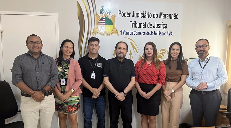 Foto horizontal de evento. Ouvidoria visita Comarca de João Lisboa. A foto mostra um grupo de sete pessoas posando lado a lado em frente a uma parede branca que possui um brasão e um texto institucional. O texto diz: "Poder Judiciário do Maranhão - Tribunal de Justiça - 1ª Vara da Comarca de João Lisboa/MA". Todos estão vestidos formalmente.
