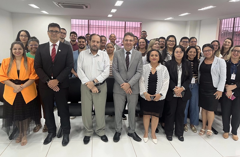 Foto horizontal de evento. A imagem mostra um grupo de pessoas posando para uma foto em um ambiente interno. A maioria está de pé, enquanto algumas pessoas estão sentadas ao fundo. Todas estão vestidas de maneira formal ou semiformal, e algumas utilizam crachás de identificação. O ambiente é bem iluminado, com luzes no teto e janelas ao fundo que permitem a entrada de luz natural.