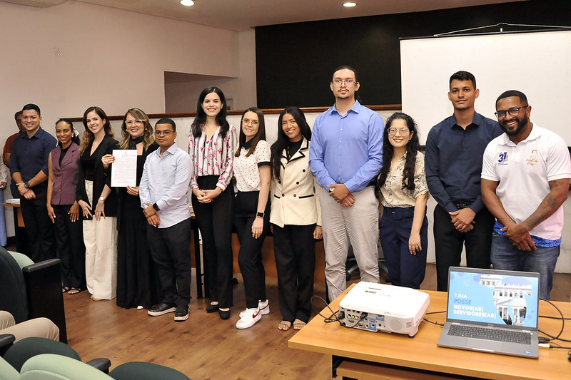 Foto horizontal da posse de novos 32 novos servidores no TJMA. A imagem mostra um grupo de treze pessoas posando para uma foto em um auditório. Todos estão vestidos de maneira formal ou semi-formal. Uma pessoa está segurando um documento, enquanto outras simplesmente posam para a câmera.  Na frente do grupo, há uma mesa sobre a qual estão um projetor e um laptop. Ao fundo, há uma tela de projeção branca e cadeiras verdes visíveis no canto esquerdo.
