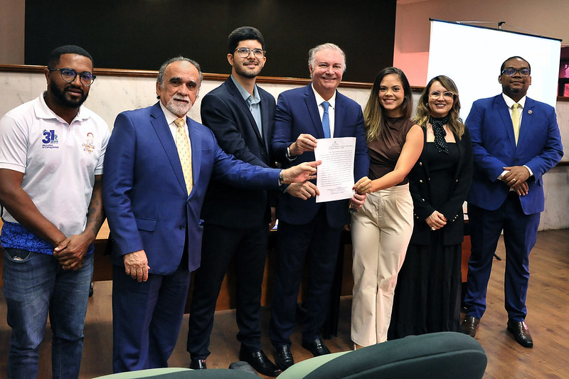 Foto horizontal da posse de novos 32 novos servidores no TJMA. A imagem mostra um grupo de sete pessoas em pé, em um ambiente de auditório. Todos estão vestidos formalmente, usando ternos e roupas sociais. No centro da foto, duas pessoas seguram um documento de termo de posse, enquanto uma delas aponta para ele, enfatizando sua relevância. Ao fundo, há uma tela de projeção branca.