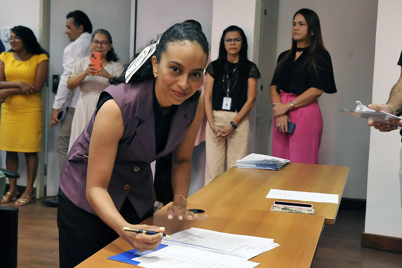 Foto horizontal da posse de novos 32 novos servidores no TJMA. A imagem mostra uma pessoa sentada diante de uma mesa enquanto assina documento. Ela está vestindo um colete roxo e uma blusa preta. Sobre a mesa estão os documentos que estão sendo assinados, além de uma caneta e um celular. Ao fundo, é possível ver várias outras pessoas, algumas em pé, observando a cena.