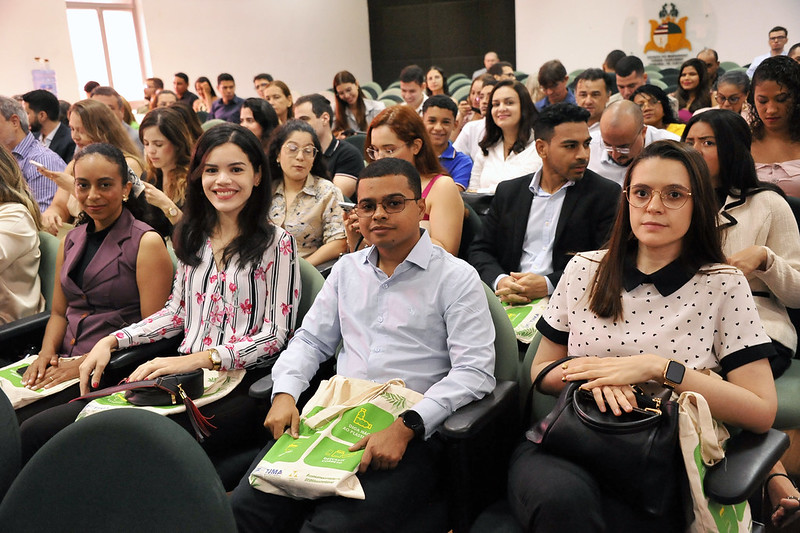 Foto horizontal da posse de novos 32 novos servidores no TJMA. A imagem mostra um grupo de treze pessoas posando para uma foto em um auditório. Todos estão vestidos de maneira formal ou semi-formal. Uma pessoa está segurando um documento, enquanto outras simplesmente posam para a câmera.  Na frente do grupo, há uma mesa sobre a qual estão um projetor e um laptop. Ao fundo, há uma tela de projeção branca e cadeiras verdes visíveis no canto esquerdo.