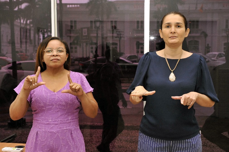Foto horizontal das servidoras do Tribunal de Justiça do Maranhão Ana Neres, à esquerda, e Gisele Viana, à direita, em frente a uma ampla janela de vidro. Ambas estão fazendo gestos com as mãos, da Língua Brasileira de Sinais. A imagem mostra as duas em pé, de frente para a câmera, em um ambiente interno. Ana, mulher negra, estatura mediana, cabelos pretos longos, usa óculos de grau com armação de metal e um vestido roxo com detalhes em relevo. Gisele, mulher branca, alta, cabelos castanhos presos na parte de trás, veste uma blusa azul escura com uma calça listrada e um colar. Ao fundo, através da janela, podem ser vistos reflexos de carros e um edifício.