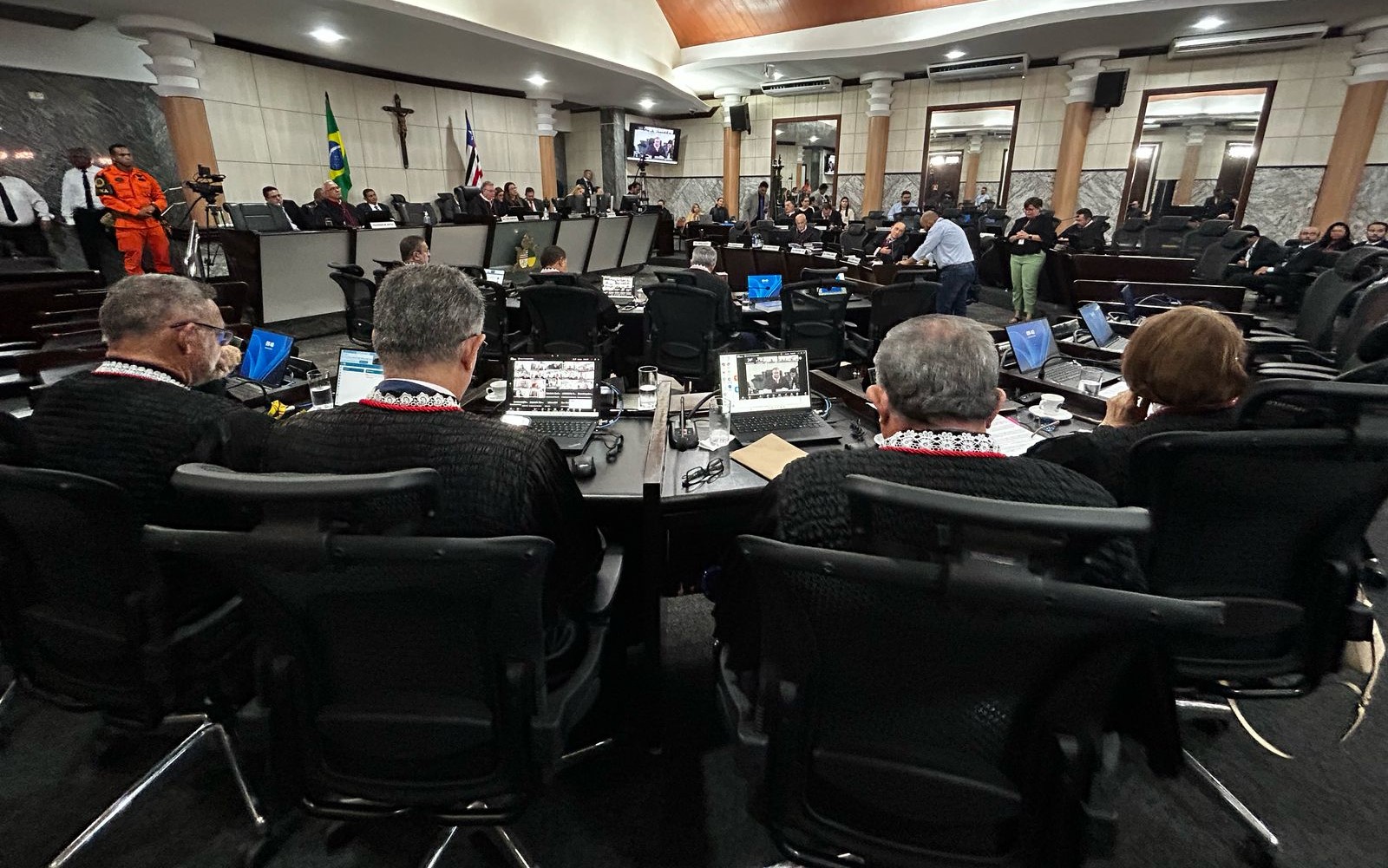 Foto horizontal da Sessão do Órgão Especial do TJMA. A imagem mostra um ambiente formal, na Sala das Sessões Plenárias do tribunal. Há várias pessoas sentadas em cadeiras, sendo que algumas estão vestindo togas pretas com detalhes em vermelho e branco, que são desembargadores e desembargadoras. Na frente das mesas, há laptops e documentos distribuídos. No fundo, é possível ver bandeiras, incluindo a do Brasil, e uma cruz na parede. Também há uma pessoa em pé vestindo um uniforme laranja, que é um bombeiro. A sala é ampla, com paredes revestidas de mármore e colunas decorativas. Câmeras de vídeo estão posicionadas para gravar o evento, e monitores exibem a sessão. A atmosfera é de seriedade e solenidade.