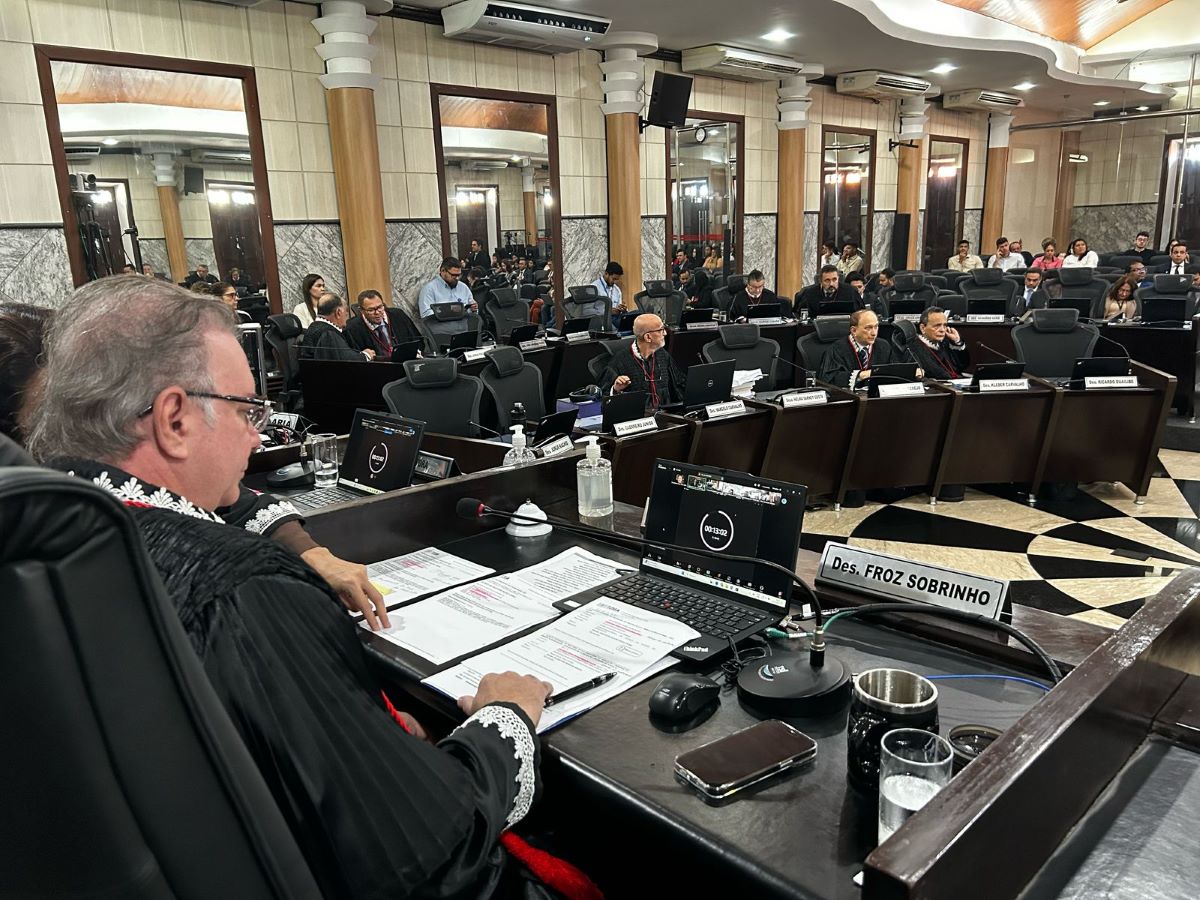 Foto horizontal da Sessão do Órgão Especial do TJMA em 12 de março de 2025.A imagem mostra uma sessão formal na Sala das Sessões Plenárias do TJMA. Há várias pessoas sentadas em cadeiras, algumas delas com laptops abertos à sua frente. Na frente da sala, há uma mesa com diversos itens: documentos, um laptop, um microfone, um copo de água e um nome identificando "Des. FROZ SOBRINHO". A cadeira neste local é ocupada pelo presidente do TJMA, desembargador Froz Sobrinho, homem branco, meia idade, cabelos grisalhos, que usa toga preta. As outras pessoas estão vestidas formalmente, algumas também com togas. O ambiente é bem iluminado, com paredes revestidas de mármore e colunas decorativas.