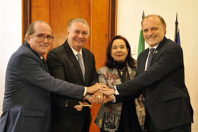 Foto horizontal de solenidade. TJMA e UFMA assinam convênio de mestrado em sustentabilidade. A imagem mostra quatro pessoas em pé - os desembargadores Ricardo Duailibe e Froz Sobrinho, a desembargadora Sônia Amaral e o reitor da UFMA, Fernando Carvalho, vestindo trajes formais. Elas têm as mãos sobrepostas no centro, como se estivessem simbolizando união ou celebrando uma parceria. Ao fundo, pode-se ver uma porta de madeira e, à direita, bandeiras estão posicionadas. Uma das pessoas está segurando um pequeno objeto dourado na mão esquerda.