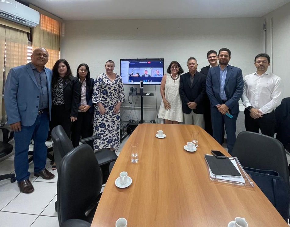A imagem mostra um grupo de nove pessoas em pé, em uma sala de conferências. Elas estão posicionadas em frente a uma mesa que possui várias xícaras de café, pires e um copo de água. Na parede atrás delas, há uma tela de televisão exibindo uma videoconferência com vários participantes visíveis na imagem. A sala tem paredes bege, uma janela com persianas e uma unidade de ar-condicionado instalada. O ambiente sugere um momento de encontro, possivelmente uma reunião presencial combinada com participação virtual.