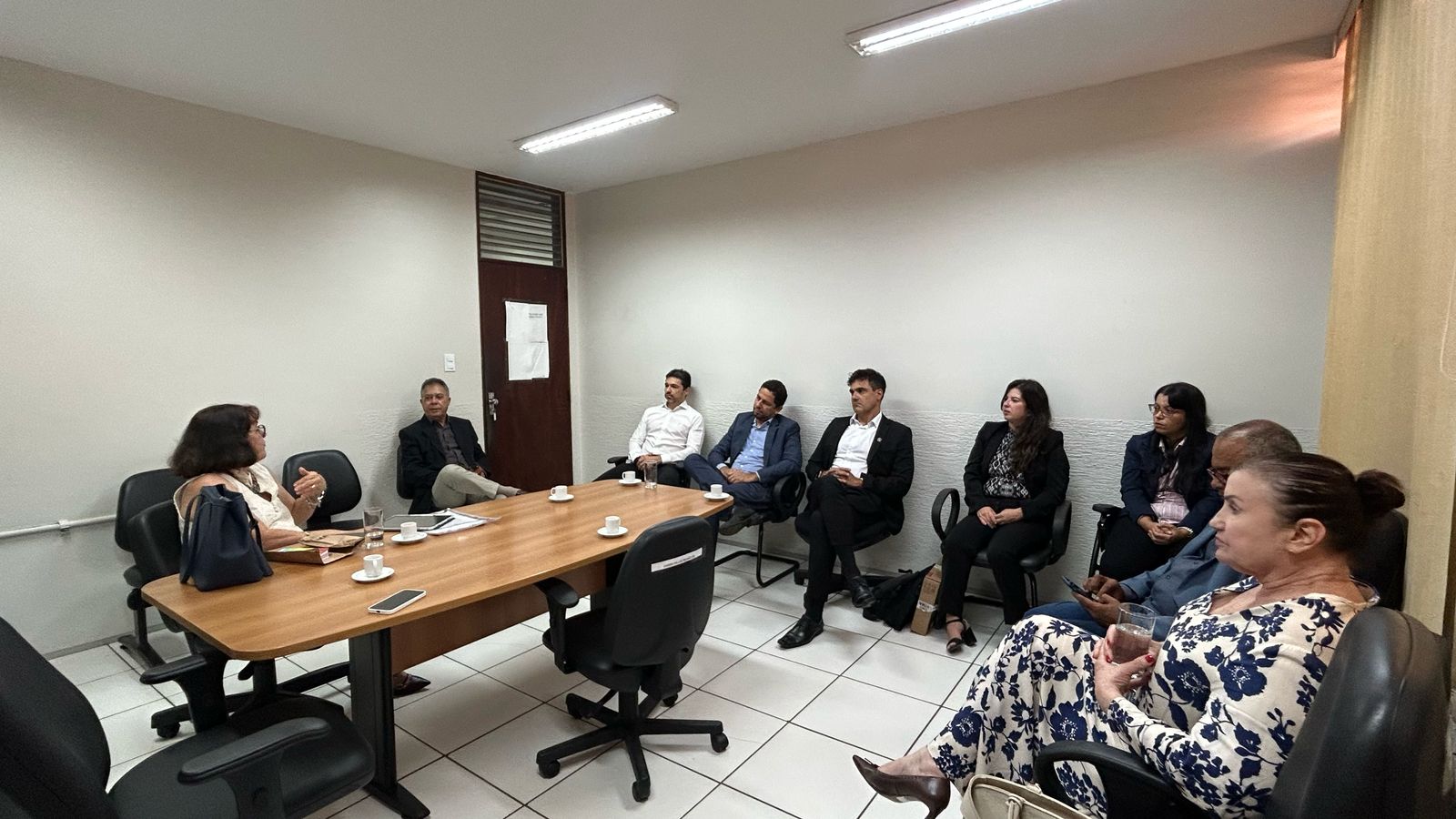 imagem mostra um ambiente de sala de reuniões com nove pessoas sentadas ao redor de uma mesa retangular de madeira. O espaço possui paredes brancas, piso de azulejo e iluminação fluorescente. Sobre a mesa, há xícaras, pires, um celular, um caderno e alguns papéis. As pessoas parecem estar engajadas em uma discussão ou reunião, demonstrando um momento profissional e colaborativo. Ao fundo, uma porta possui um papel fixado, mas o texto nele não é legível. Esse cenário reflete um ambiente de trabalho ou organizacional, onde questões importantes estão sendo discutidas