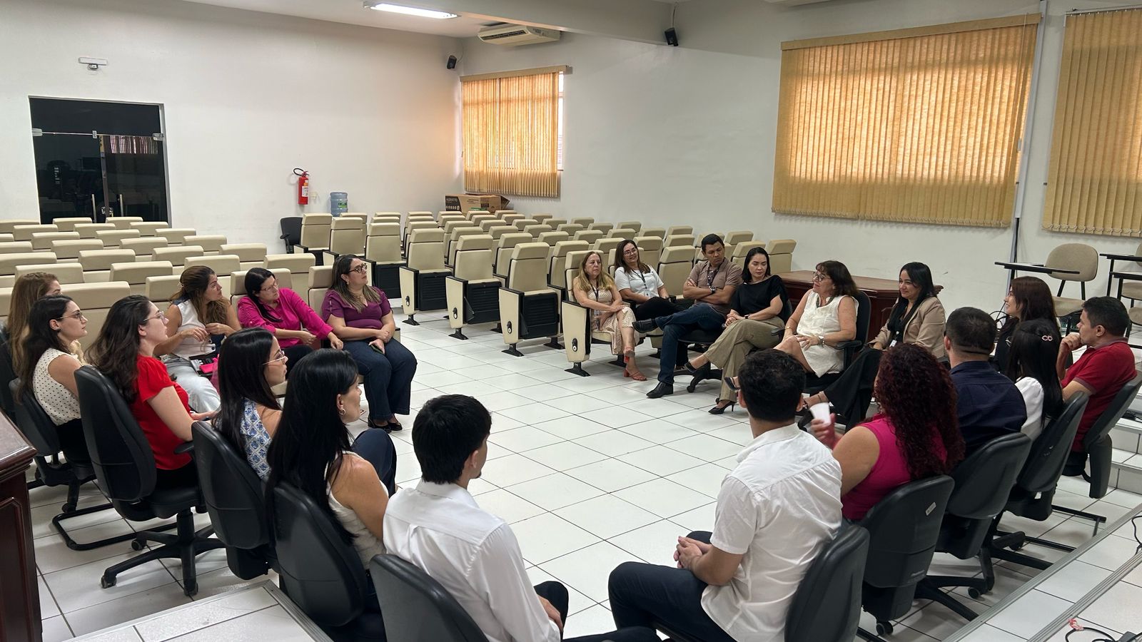 A imagem mostra um grupo de pessoas sentadas em círculo, participando de uma reunião ou discussão. O ambiente parece ser uma sala de conferências ou de treinamento, com várias cadeiras bege organizadas em fileiras que estão desocupadas. Há um extintor de incêndio preso à parede e uma janela com persianas fechadas. As pessoas estão sentadas em cadeiras de escritório