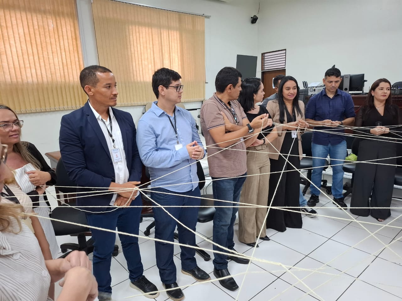 imagem mostra um grupo de pessoas em pé, formando um círculo e segurando pedaços de barbante que estão conectados entre si, formando uma estrutura semelhante a uma teia. Essa atividade parece ser um exercício de construção de equipe, provavelmente com o objetivo de promover a comunicação, a colaboração e a confiança entre os participantes. O ambiente é uma sala interna, com cadeiras de escritório e mesas ao fundo, sugerindo que a atividade está ocorrendo em um ambiente de trabalho ou de treinamento. Os barbantes conectando as pessoas simbolizam interconexão e trabalho em equipe, tornando o exercício visualmente e simbolicamente interessante para fortalecer a dinâmica de grupo