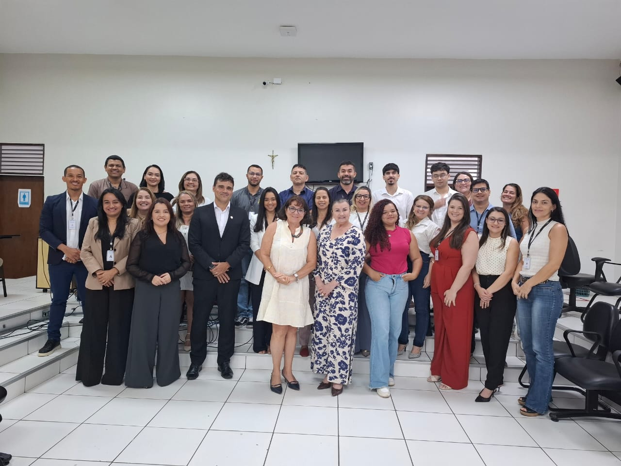 A imagem apresenta um grupo de aproximadamente 23 pessoas reunidas em um ambiente formal ou profissional, como uma sala de reuniões ou conferências. A maioria está vestida com roupas de trabalho ou semiformais. No fundo da sala, há uma parede branca com um crucifixo e um pequeno quadro negro ou tela. Também é possível ver cadeiras e uma mesa no ambiente. O grupo parece estar junto para um evento, reunião ou foto em grupo, representando uma equipe ou organização